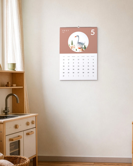 Calendrier accroché au mur au-dessus d'une cuisine jouet en bois d'enfant, ouvert sur mai : une oie en papier plié sur fond terracotta.