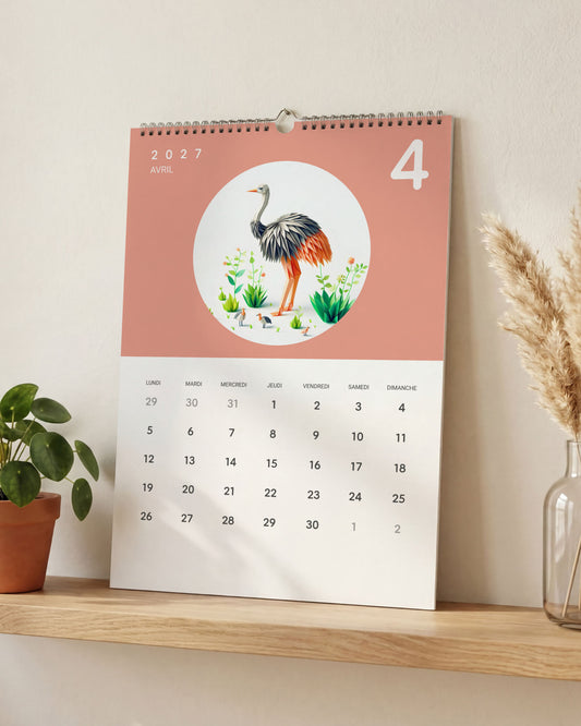 Calendrier posé sur une étagère en bois clair, ouvert sur avril : une autruche en papier plié sur fond rose saumon, entre une plante verte et un vase d'herbe de la pampa.
