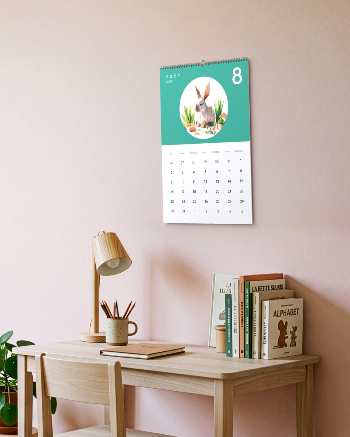 Calendrier accroché au-dessus d'un bureau d'enfant en bois, à côté d'une lampe et d'une pile de livres, ouvert sur août : un lapin en papier plié sur fond sarcelle.