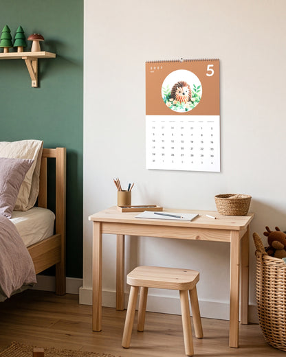 Calendrier accroché au-dessus d'un petit bureau d'enfant en bois, près d'un tabouret et d'un panier en osier, ouvert sur mai : un hérisson en papier plié sur fond terracotta.