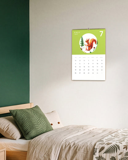 Calendrier accroché au mur vert d'une chambre d'enfant, au-dessus d'un lit aux coussins beige et vert, ouvert sur juillet : un écureuil en papier plié sur fond vert clair.