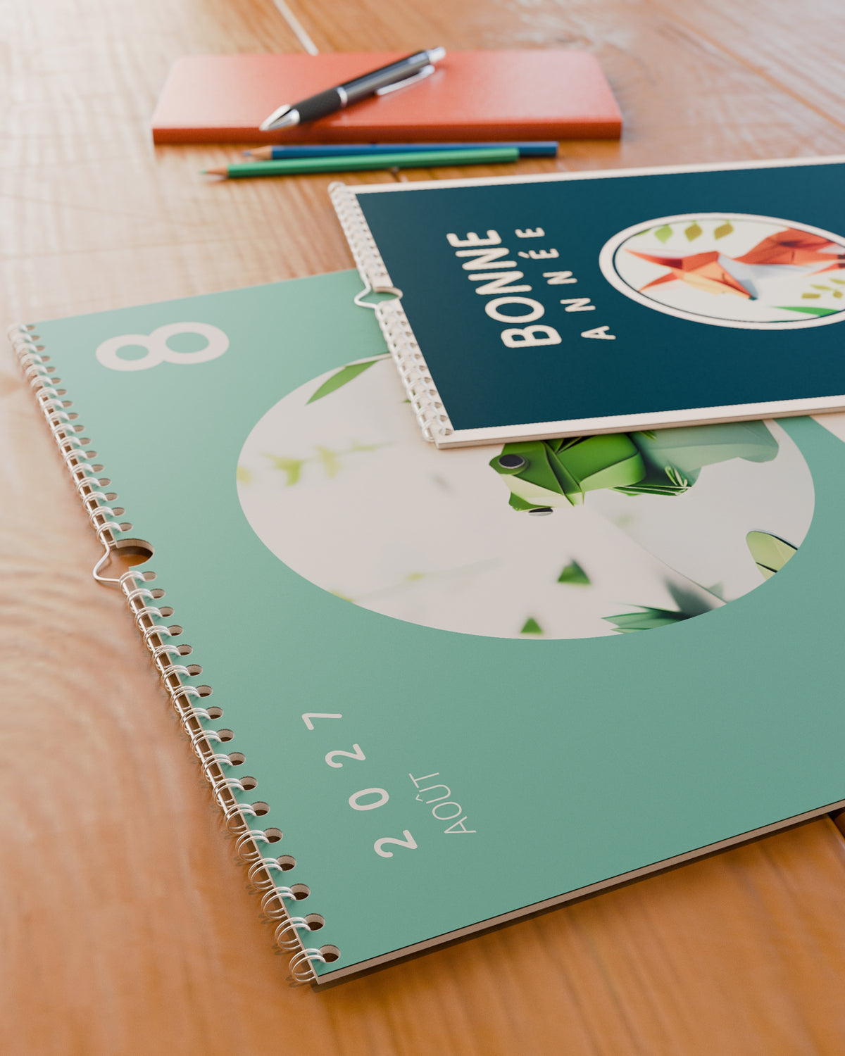 Calendrier mural Bonne Année 2027 posé à plat sur un bureau en bois, ouvert sur août : une grenouille en papier plié sur fond vert d'eau. La reliure spirale et la couverture bleu canard au renard sont visibles.