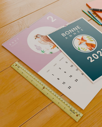 Calendrier mural Bonne Année 2027 posé sur un bureau à côté d'une règle de 30 cm, pour donner un repère d'échelle. Il est ouvert sur février : un rapace en papier plié sur fond mauve.
