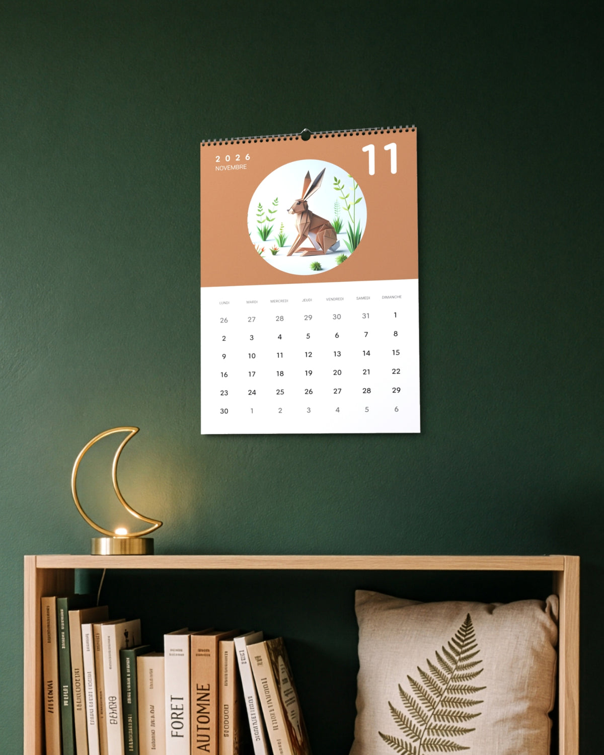 Calendrier accroché au mur vert sapin au-dessus d'une étagère avec livres et lampe en forme de croissant de lune, ouvert sur novembre : un lièvre en papier plié sur fond terracotta.