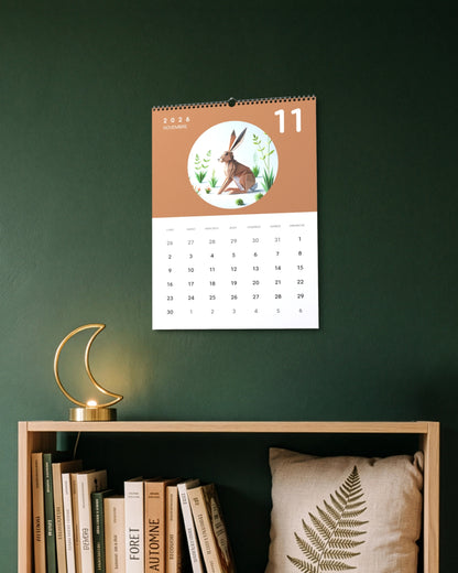 Calendrier accroché au mur vert sapin au-dessus d'une étagère avec livres et lampe en forme de croissant de lune, ouvert sur novembre : un lièvre en papier plié sur fond terracotta.
