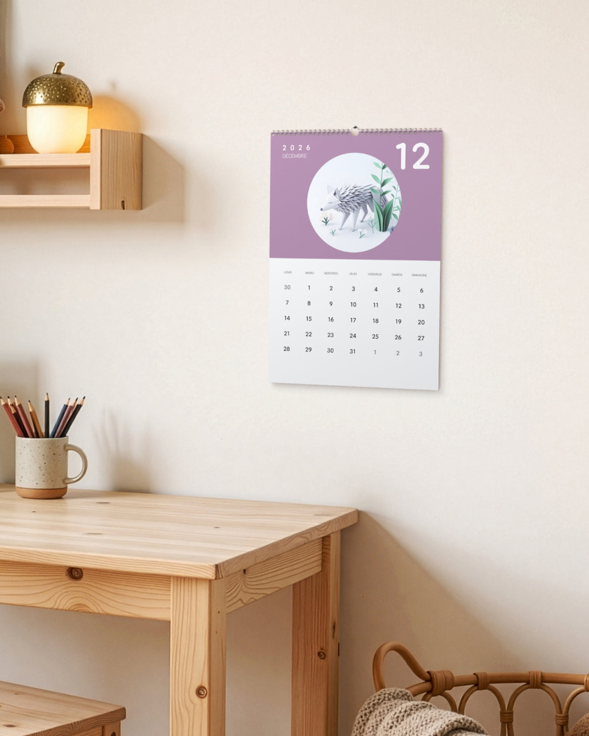 Calendrier accroché au-dessus d'un bureau d'enfant en bois, à côté d'une lampe en forme de gland et d'un mug à crayons, ouvert sur décembre : un loup en papier plié sur fond mauve.