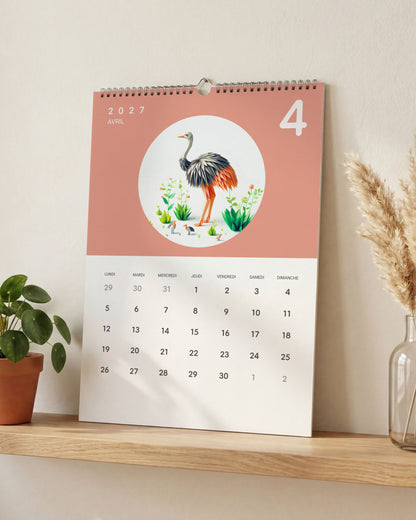 Calendrier posé sur une étagère en bois clair, ouvert sur avril : une autruche en papier plié sur fond rose saumon, entre une plante verte et un vase d'herbe de la pampa.