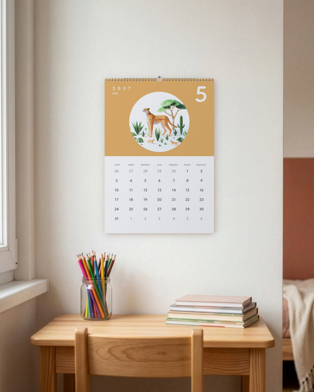Calendrier accroché au-dessus d'un petit bureau d'enfant près d'une fenêtre, à côté d'un bocal de crayons de couleur et d'une pile de cahiers, ouvert sur mai : un guépard et son petit en papier plié sur fond ocre.