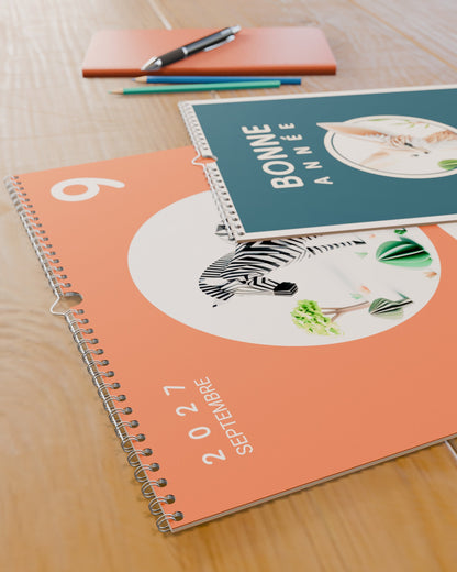 Gros plan sur la reliure spirale et le grain du papier du calendrier mural Bonne Année 2027, posé sur un bureau d'enfant.