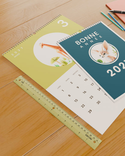 Calendrier mural Bonne Année 2027 posé sur un bureau à côté d'une règle de 30 cm, pour donner un repère d'échelle.