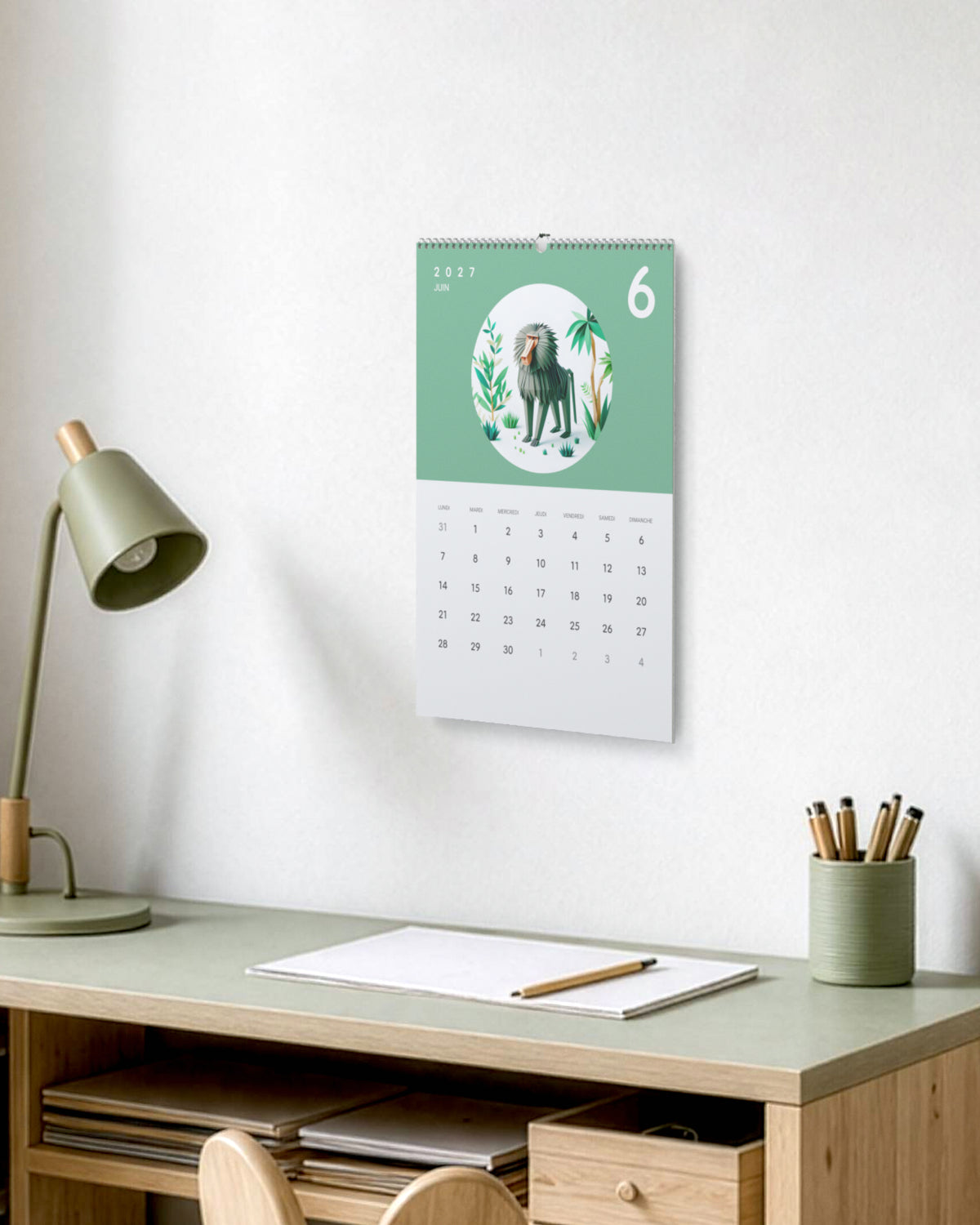 Calendrier accroché au-dessus d'un bureau d'enfant en bois clair, à côté d'une lampe verte et d'un pot à crayons, ouvert sur juin : un babouin en papier plié sur fond vert.