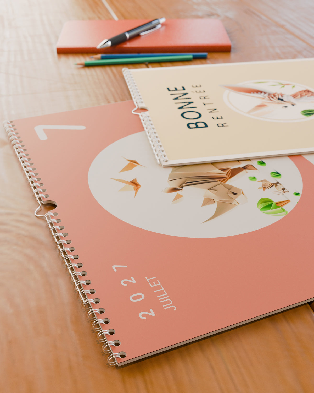 Calendrier mural Bonne Rentrée 2026 / 2027 posé à plat sur un bureau en bois, ouvert sur juillet : une autruche en papier plié sur fond corail. La reliure spirale et la couverture crème au fennec sont visibles.